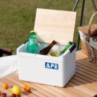 A white cooler with a wooden lid labeled APE is open, revealing various bottled drinks inside. It sits on a table outdoors, surrounded by fruit and near a wicker basket and grass lawn.