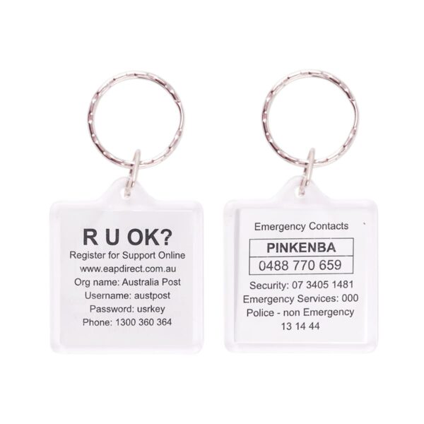 A keychain with two key tags. The left tag prompts R U OK? with registration info for a site. The right tag lists emergency contacts for Pinkenba, including phone numbers for security and services.