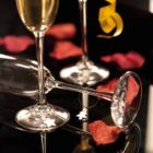 Two champagne flutes, one upright and one tipped over, are surrounded by red rose petals on a reflective black surface. A small tree-shaped charm dangles from the base of the tipped flute, and a yellow ribbon is blurred in the background.