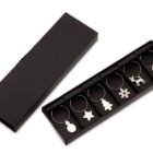 A set of six silver wine glass charms with holiday-themed shapes: round ornament, star, Christmas tree, snowflake, reindeer, and stocking. They are neatly arranged in an open black box.