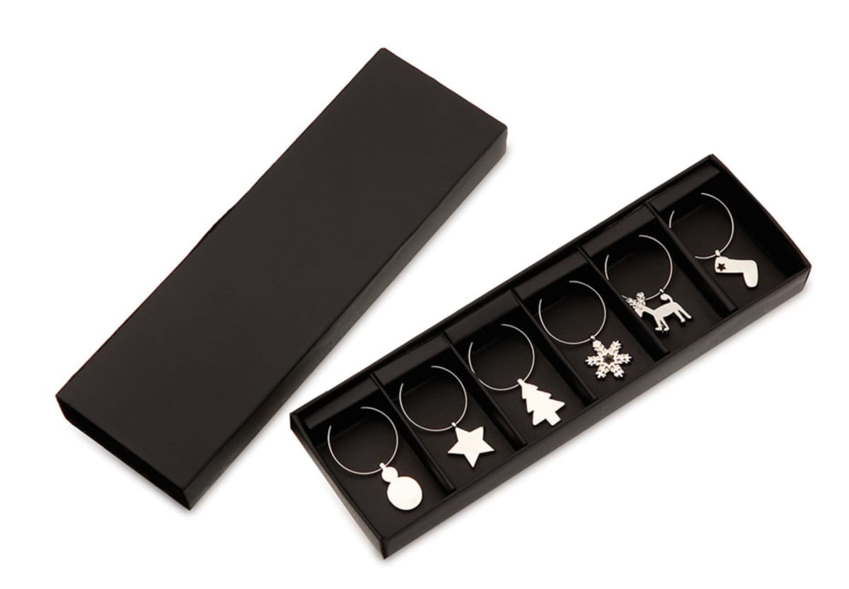 A set of six silver wine glass charms with holiday-themed shapes: round ornament, star, Christmas tree, snowflake, reindeer, and stocking. They are neatly arranged in an open black box.