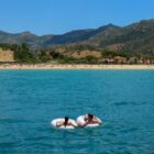 A person relaxes on an inflatable ring in calm turquoise waters, facing a sandy beach with several people. The background features rolling green hills and a clear blue sky.