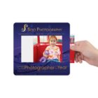 A photo frame from Soo Photography with a hand holding a picture of a child sitting in a red seat, wearing a tie-dye shirt and holding a purple stuffed animal. The frame reads captured by the Photographer of the Year.