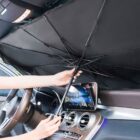 A person is setting up a black foldable umbrella-style sunshade inside a car, attaching it to the windshield. The rearview mirror and part of the steering wheel are visible.