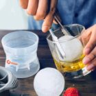 Close-up of a hand using tongs to place a spherical ice cube into a glass of amber liquid. An empty ice mold and another ice sphere are on a wooden table, with a strawberry nearby.