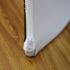 Close-up of a white zipper on a leather or fabric item, partially open, with a brown edge. The background is a wooden floor, providing contrast to the bright material and the metallic zipper.