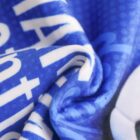 A close-up of a blue fabric with white text and patterns, slightly blurred to create a soft texture. The fabric appears to be gathered or crumpled in the center, casting gentle shadows.