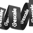 A black ribbon with the word escaler and a floral logo printed in white repeatedly along its length. The ribbon is partially coiled with a textured surface.