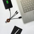 A multi-port charging cable is connected to a laptop and charging a smartphone. The phone displays a charging icon. A tablet and a stylus are partially visible nearby on a white table.