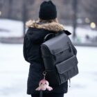 A person with long hair is wearing a black winter coat with a fur-lined hood and a black beanie. They have a large black backpack with a cute, pink plush toy keychain attached. The background shows a snowy park.