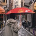 A person in a khaki coat walks down a cobblestone street holding a red umbrella with floral designs. The street is lined with colorful buildings, and the sky appears overcast.