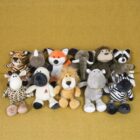 A collection of stuffed animals is arranged in two rows on a yellow background. The toys include a lion, giraffe, zebra, elephant, fox, monkey, raccoon, tiger, and hippo, featuring various colors and patterns.