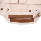Close-up of a beige canvas bag with brown leather handles and metal rivets. The handles are stitched to the bag, and the fabric features a textured pattern.