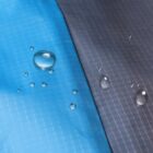 Close-up of a blue and dark gray waterproof fabric with water droplets beading on the surface, demonstrating its water-resistant properties. The fabric has a grid-like texture.