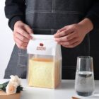 A person in a dark apron sealing a clear plastic bag with a slice of thick, square bread labeled Homemade Bakery. A glass of water and a white rose are on the white table next to the bag.