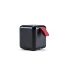 A small, black, cube-shaped speaker with rounded edges is shown. It features a red fabric strap on one side and a micro USB charging port. The design is minimalist, set against a plain white background.