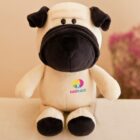 A plush toy dog with cream-colored body and black ears, snout, and paws sits on a surface. Its paws have hearts, and kidSPLACE is printed on its chest. A blurred pink flower arrangement is in the background.