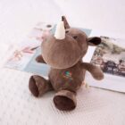 A cute brown plush rhinoceros toy with a white horn sits on a textured white surface. Behind it, there is an open magazine featuring colorful images. The plush toy has stitched details and a playful, soft appearance.