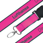 A pink lanyard with the word Lufthansa in gray text. It features a black plastic buckle and a metal clip at the end. The edges of the lanyard are dark green.