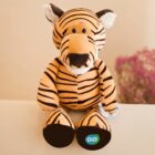 Plush tiger toy sitting upright on a beige surface. It has black stripes and features, with ears tipped in white. The paws are marked with a green and blue logo featuring the word GO. In the background, there are pink and white flowers.
