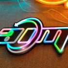 A colorful neon sign on a textured surface displays the stylized letters ADM with a swoosh design around them, featuring bright shades of pink, blue, green, and orange.