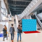 Four people stand inside a modern art gallery with a red carpet and blue graphic wall displaying Art World text. The space features large sculptures, an escalator, and stairs, all under a high ceiling.