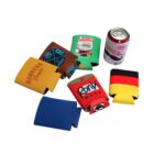 Various colorful can koozies with different logos and designs are laid out around an unopened can of Pabst Blue Ribbon beer. The designs include company logos, abstract patterns, and national flag colors.