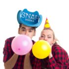 Two people in party hats blowing pink and yellow balloons, wearing plaid shirts.