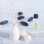 A white ceramic vase with reed diffusers, two decorative rattan balls, black stones, and a small bottle of yellow-green essential oil are arranged on a light wooden surface.