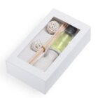 A white box with a transparent lid containing a small bottle of yellow liquid, wooden sticks, a white vase, and two decorative white spherical objects.