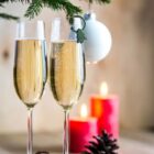 Two champagne flutes filled with bubbly liquid sit beneath a small pine branch adorned with a white ornament. In the background, three lit red candles and a pine cone create a cozy atmosphere.
