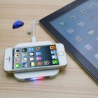 A smartphone with a visible home screen is placed on a wireless charger, emitting a red and blue light. A tablet with an app-filled screen is nearby on a wooden surface, alongside a blue paper weight and a small, round decorative item.