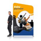 A man in a suit stands next to a colorful promotional banner. The banner features the text Enjoy your travel and a photo of three people with backpacks, sitting on rocks, raising their arms towards a scenic view. Additional text is included below the image.