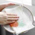 A person washing a folded, ribbed cloth with a red and green design under a stream of water in a sink. The cloth is placed on a white plate with a brown rim. Hands are applying gentle pressure to clean it.