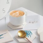 A cup of latte with a leaf-patterned foam design on a hexagonal coaster, next to a gold spoon. A piece of paper with a floral illustration appears beneath the coaster. The background includes the edge of a clock face and additional coasters.