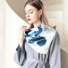 A woman with long hair wears a gray dress and a blue patterned scarf. She is standing indoors, touching the scarf gently while looking downward. The background features white, curved architecture.
