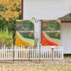 Two decorative garden flags with Happy Thanksgiving text, featuring autumn leaves and pumpkins. One flag has a green theme, the other orange. They stand in front of a white picket fence and leafy trees, with part of a house visible.