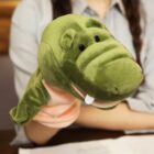A person with braided hair holds a green plush alligator hand puppet with white teeth. The puppets mouth is open slightly, and the person is wearing a grey shirt. Papers are visible on the table in front of them.