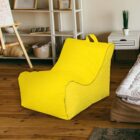 Yellow bean bag chair on a wooden floor in a cozy room. It is situated near a white dresser and a shelf with various decorative items. An easel is visible to the left, and part of a bed with a white comforter is on the right.