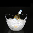 A clear ice bucket filled with ice holds a single bottle of beer. The bottle is angled slightly with the label partially visible. The bucket sits on a dark, reflective surface.