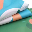 Close-up of folded fabric with abstract patterns. The top layer features blue, white, and peach colors, while the bottom layer shows green shades. The texture appears smooth and slightly glossy.