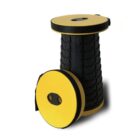 A black collapsible stool with yellow round seats is partially extended. Another similar seat with two oval holes is leaning against it. The background is plain white, highlighting the stools compact design.