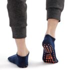 Two feet wearing dark blue grip socks with orange patterns on the soles, standing on a white surface. The person is wearing dark gray pants.