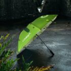 A bright green umbrella with the word CUMBRA on it lies open on a wet, dark pavement. It is surrounded by green plants, with a textured, dark wall in the background. The setting suggests a rainy environment.