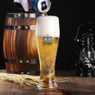 A glass of beer is being poured with a frothy head. The glass sits on a wooden table beside wheat stalks. In the background, theres a wooden barrel and an old-fashioned lantern. The glass has Premium Quality written with two clinking mugs.