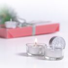Two small glass candles, one lit and one unlit, sit on a white surface. In the background, theres a red gift box with a silver ribbon, and some greenery, slightly out of focus.