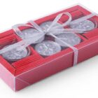 A rectangular gift box with a clear lid, containing six silver tealight candles arranged in pairs. The box is decorated with a shiny silver ribbon tied in a bow. The sides of the box are red with a striped texture.