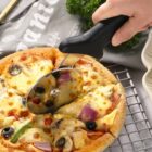 A hand using a pizza cutter to slice a freshly baked pizza topped with olives, mushrooms, onions, and bell peppers. The pizza is on a cooling rack, and a sprig of parsley is visible in the background.