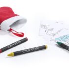 A red and white plush Christmas stocking with three crayons—red, yellow, and green—next to it. Several coloring sheets feature outlines of a gift box, star, and decorated Christmas tree.