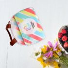 A colorful lunch bag with diagonal stripes is placed on a white surface. The bag has brown straps and a central logo placeholder. Beside it, there is a bowl of mixed berries and a small bouquet of vibrant flowers.